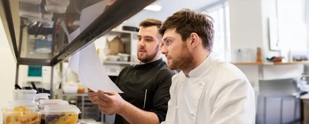Köche in der Küche besprechen ein Rezept oder Bestellzettel, konzentriert und im Gespräch vertieft.