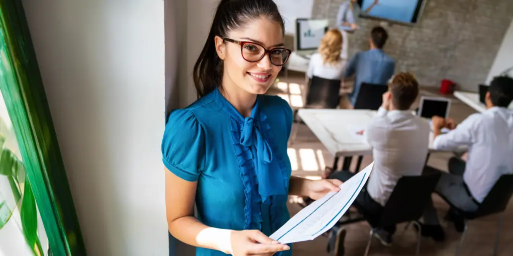 Geschäftsfrau präsentiert lächelnd in einem modernen Büro, während hinter ihr ein Meeting stattfindet. Büro, Teamwork.
