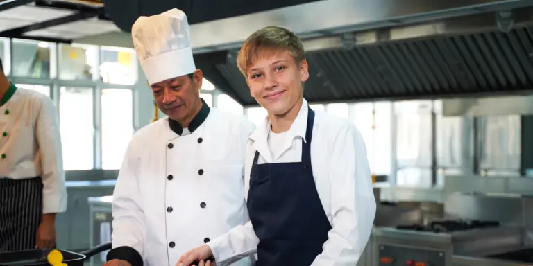 Zwei Köche, ein Erwachsener und ein Junge, lächeln in einer modernen Restaurantküche.