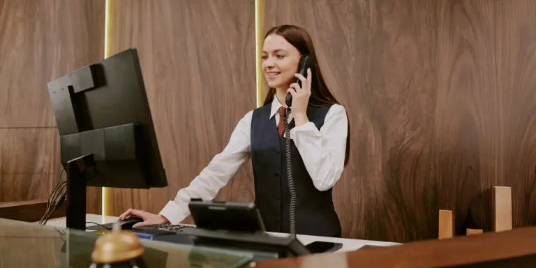 Eine lächelnde Rezeptionistin telefoniert an einem Empfangstresen mit Computer.