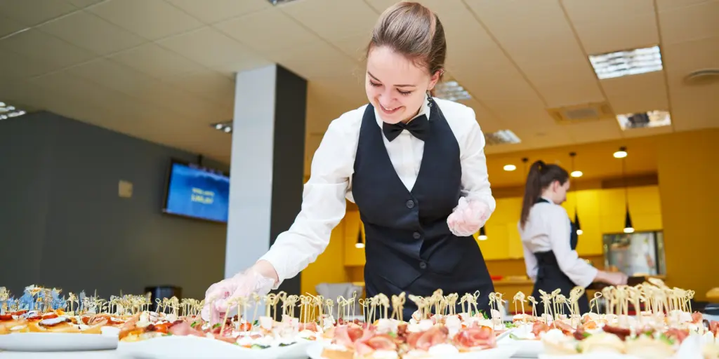Kellnerin richtet sorgfältig Snacks auf einem Tisch an, elegant gekleidet in Schwarz-Weiß-Kleidung.